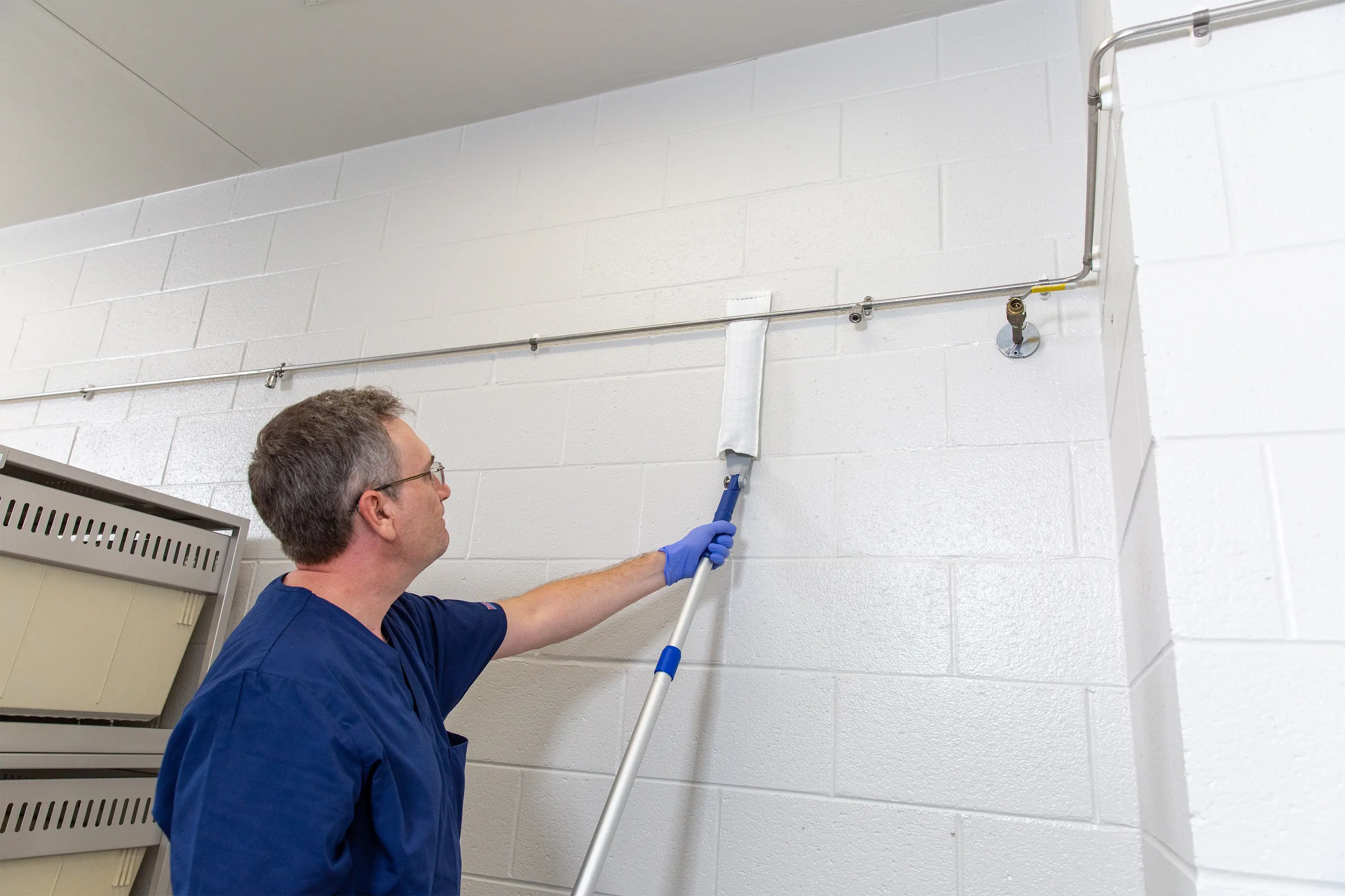 Worker using an extendable high duster to clean a wall in a controlled facility.